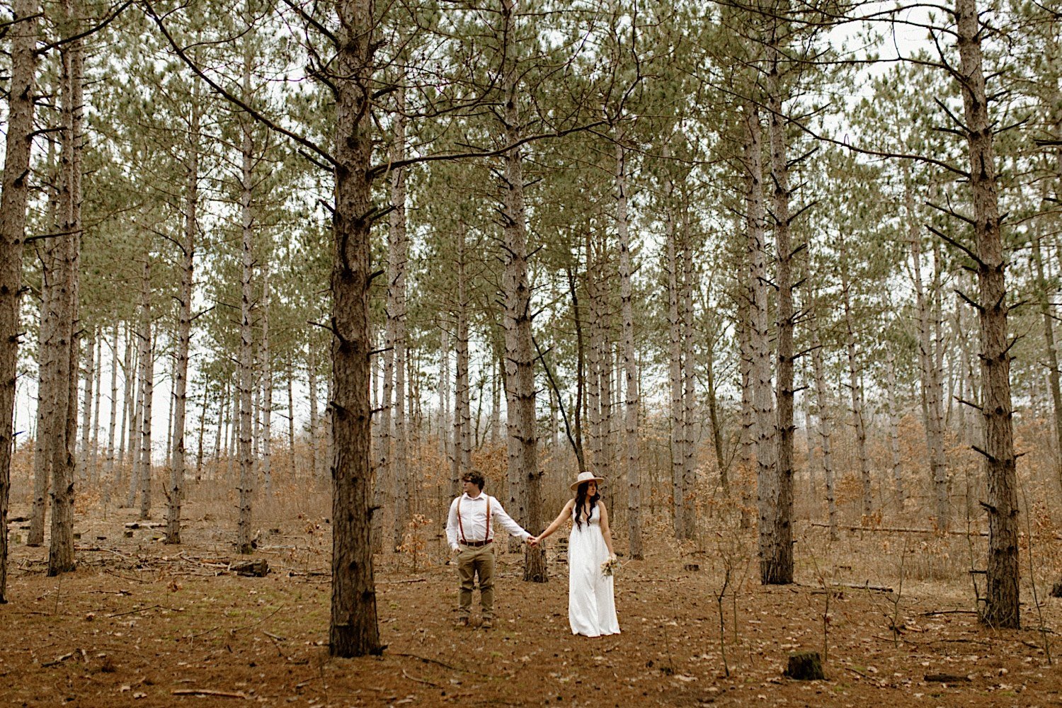 A man in a white shirt and khaki pants and a woman in a white dress and hat stand in a forest facing the camera holding hands and looking away from each other.